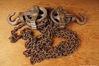 Two metal hoist pulleys with large hooks attached and several feet of chain laid out on a wooden surface.