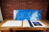 Blue acrylic sheets of multiple sizes arranged in row with protective blue covering visible against brick wall background, white artist materials in front on a wooden surface.