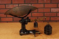 Front angle view of the antique cast iron merchant's scale showing the brass weighing pan, the black painted cast iron base, the brass balance beam and one weight hanging.