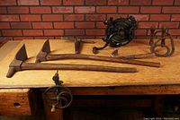 Overall lot on wooden bench showing three pick mattocks, cast iron multi-handle tool, two wire mashers, T-handle gimlet and bench crank clamped to edge