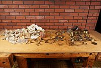 Wide shot showing many brass lamp parts and cloth wicks laid out on wooden table against brick wall background.