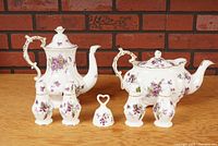 Two porcelain teapots with matching floral decorations, two pairs of salt and pepper shakers, and a small porcelain bell arranged on a wooden surface against a brick wall background.