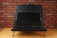 Front view of the trapezoidal black metal wood stove fireplace resting on angled legs against a brick wall background.