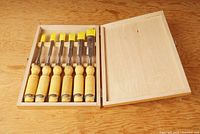 View of the six chisels neatly arranged inside an open wooden case on a wooden surface. Shows the full set and the storage case lid open.