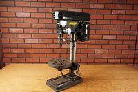 Side angled view of the drill press showing metal base, round adjustable table, motor housing, and chuck with drill bit.