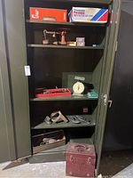 Garage cabinet with multiple shelves showing various items including boxed filters, old scale, tools in red tray, and metal andiron pieces on lower shelves.