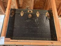 Front view of large antique black trunk showing metal lock latch, corner reinforcements, and scuff marks.