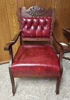 Front view of antique Victorian chair showing red button-tufted leather seat and backrest and carved wood top detail.