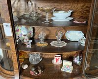 Photo showing bottom three shelves inside curio cabinet with a variety of glassware and decorative ceramics including clear glass compote dish, pink Depression glass candle holders, and various porcelain and ceramic pieces.