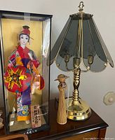 Glass-encased Asian doll in traditional colorful clothing, brass lamp with etched glass panels, and small wooden figurine on a piano surface.
