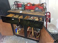 Overall front view of black wooden workbench with drawers open showing tools inside, extension cords, vintage jar and chargers on top.