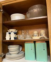 Photo showing mid century dishware including white stacked plates and bowls, vintage ceramic salt and pepper shakers shaped like figures, small glass pitchers, blue heart pattern mugs, and light blue metal tins labeled flour and sugar with copper tops.