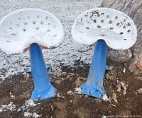 Front view showing pair of tractor seat stools with blue bases and white perforated metal seats, some rust and paint chipping visible.