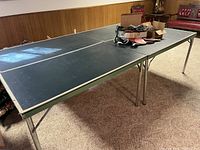 Photo of vintage ping pong table from an angle showing full table and legs, placed in a basement room with carpet, wood panel walls, and vintage couch.