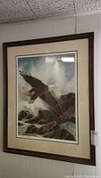 Framed wall art showing a bald eagle flying over rocky shores, with matting and dark wood frame seen against a wall.