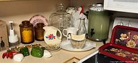 Wide view of the countertop showing most items including glass domed cake plate, amber canisters, ceramic pitcher, pepper and salt shakers, glass bottle, beverage dispenser, and trays.