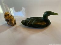 Photo shows the two figurines side by side: green stone bird with smooth polished finish, and a small ivory-colored carved figure with Asian style clothing.