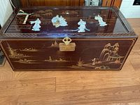 Front view of the rectangular dark brown lacquered wooden trunk showing the metal locking mechanism and mother-of-pearl inlaid figures and painted scenery.