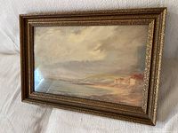 Front angled view of the coastal oil painting in a gold frame showing the ocean, beach, buildings, and cloudy sky.