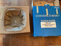 Square clear glass lid on top of original cardboard insert with blue Corning Ware box alongside.