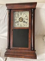 Front view of the wooden mantle clock showing the dial with Roman numerals and floral motifs, wooden frame with columns and glass door.