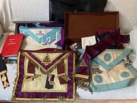 Overview of various Masonic aprons, collars, medals, and accessories displayed on a white couch including a red book and a small box with pins.