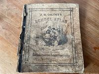 Front cover of J.H. Colton's School Atlas showing wear, discoloration, and black spine binding.