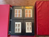 Photo of one open red binder showing black album pages with blocks of four unused stamps featuring landscapes and animals.