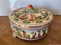 Side and top angle showing detailed cherub reliefs, floral decorations, and lid with fruit-shaped handle