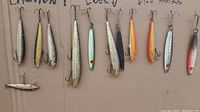 Photo of 12 vintage fish hook lures laid out on cardboard, showing various colors and shapes, with treble hooks attached