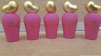 Five pink mini perfume bottles with gold heart-shaped caps, showing front sides with gold cursive writing on each bottle.