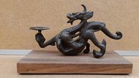 Full side view of the metal dragon statue with raised claw holding a small circular candle holder, mounted on a wooden base.