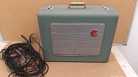 Closed view of green rectangular travel case with silver speaker grille and red RCA logo on front, carrying handle and metal clasps.