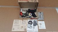 Photo showing older model Weller soldering gun with pistol grip, original box, and printed manual and soldering tips guide spread out around it, along with accessories including a tube of grease and rubber bulb.