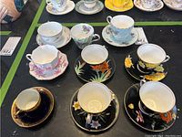 Teacups and saucers arranged showing various floral hand-painted designs and colors including black base with orange and yellow flowers, pastel blue with gold trim, and others.