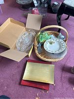 Wide shot of lot showing wicker basket with plates, Bohemian crystal oval dish in box, brass and red serving trays on floor near basket.