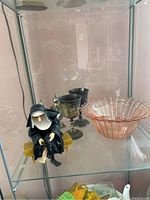 Photo of nun figurine sitting on small bench with two metal chalices and pink glass bowl behind.