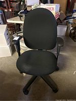Front view of black rolling computer chair with padded seat and back, adjustable armrests, situated in a cluttered room.