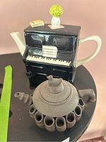 Overall view showing two ceramic teapots: piano-shaped with decorations and dark brown teapot with unique design