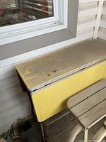 Side view of the folding side table showing the yellow melamine top and rusted metal frame and legs with some dirt accumulated on surface.