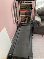 Front and running belt of Free Spirit folding treadmill placed near bookshelf and chair indoors.