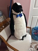 Large stuffed Emperor penguin shown sitting on a chair, showcasing front and side views with blue bow tie visible, details of fabric texture and some stains.