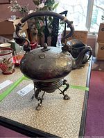 Front and side angle of vintage silver plate teapot with detailed floral engravings, mounted on candlewick stand with claw feet.