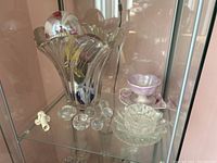 Wide shot showing a clear glass fluted bowl or vase with colored interior, multiple spoon rests including a pink and white ruffled cup, and other glassware on a glass shelf.