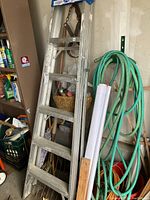 Full view of 6 ft aluminum stepladder leaning against wall with garden hose and tools in background.