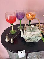 Three hand-painted wine glasses, ceramic teapot with floral design and woven handle, and two candle extinguishers on a black surface.