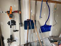 Wide shot of tools hanging and leaning against wall including Worx 18V trimmer and garden tools