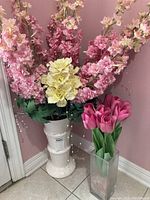 Tall white ceramic vase with pink and yellow faux flowers alongside clear rectangular glass vase with pink tulip faux flowers, displayed against a pink wall.