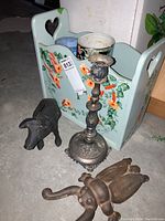 Photo showing hand-painted magazine rack, metal candlestick holder, cast iron boot puller, and wooden elephant figurine arranged on floor.