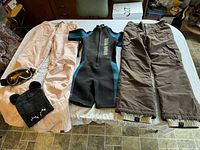 Full lot view showing two pairs of pants, wetsuit, goggles, and toque laid out on a bed.
